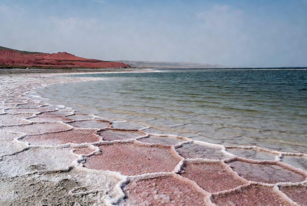Close view of Permian Zechstein evaporite formations along the sea shore, showing intricate white and pink salt crystals, gypsum, and salt polygons on mudflats with the turquoise sea and red sedimentary hills in the background.