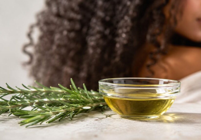 Rosemary essential oil for hair beside fresh rosemary, botanical oil, and healthy textured hair.